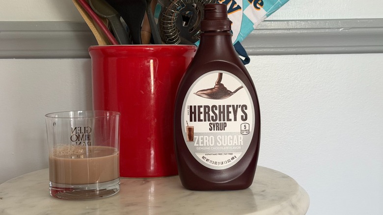 A bottle of Hershey's zero sugar chocolate syrup next to a glass of chocolate milk