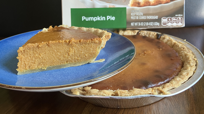 A slice of  Marie Callender's Pumpkin Pie on a blue plate sits atop a burned and crumbling pumpkin pie in a misshapen tin