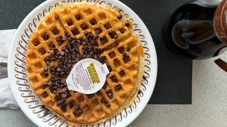 A chocolate chip waffle on a Waffle House table on a white plate with a container of whipped spread on top and a bottle of syrup to the right of it