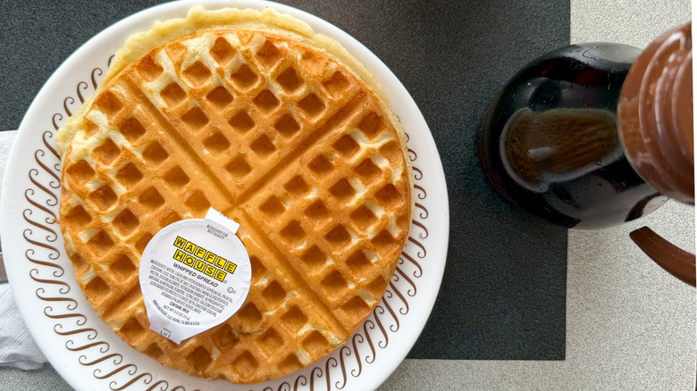 A classic waffle on a Waffle House table on a white plate with a container of whipped spread on top of it and a bottle of syrup to the right of the plate