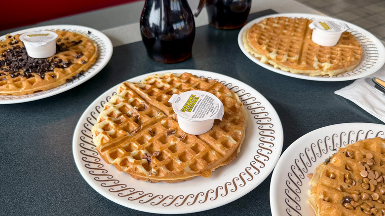 Several waffles on a Waffle House table on white plates with whipped spread containers on top of each and a pitcher of syrup in the background