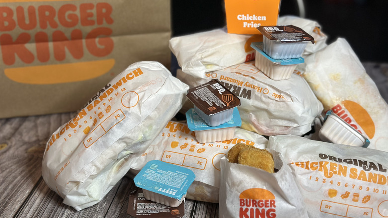 Pile of Butrger King Chicken menu items in wrappers on table, including original chicken sandwich, chicken nuggets, honey mustard chicken wrap, and many sauce packets