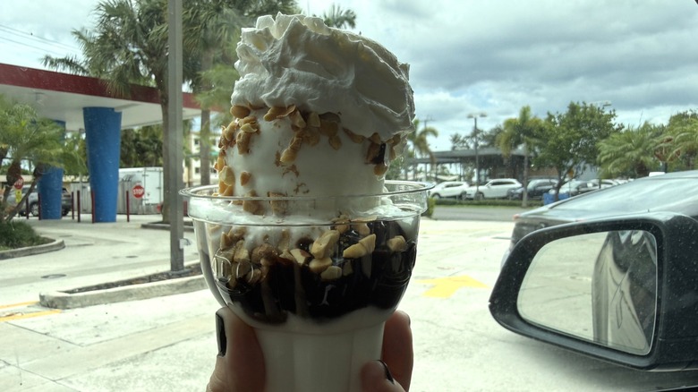 Dairy Queen hot fudge sundae topped with nuts and whipped cream held out car window in DQ parking lot