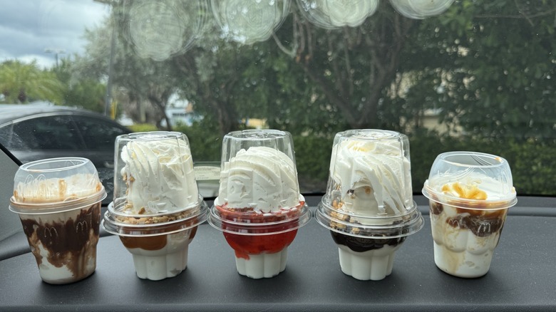 Multiple vanilla sundaes in takeout cups on car dashboard, including hot fudge from McDonald's and caramel from Dairy Queen