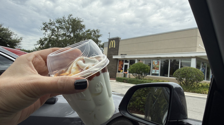hand holding hot caramel sundae in takeout cup from Mcdonald's out the car window in McDonald's location parking lot