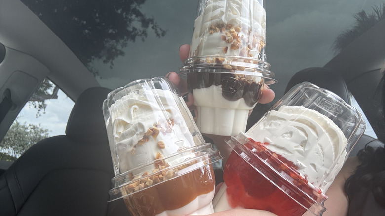Three ice cream sundaes from Dairy Queen in takeout cups held inside car, including hot fudge, caramel, and strawberry flavors topped with whipped cream and nuts.