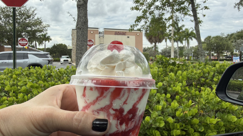 hand holding a Sonic Strawberry sundae topped with cherry in takeout cup out the car window with Sonic location in background.
