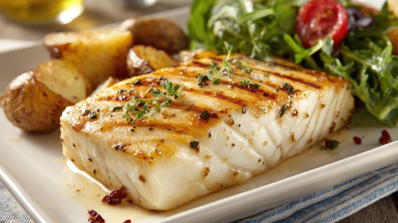 Close up of a seared cod fillet with roasted potatoes and a green salad in the background