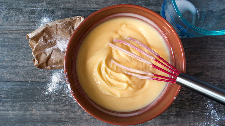 Brown bowl with lemon pudding mixed with a whisk, instant pudding powder, and a glass measuring cup with milk