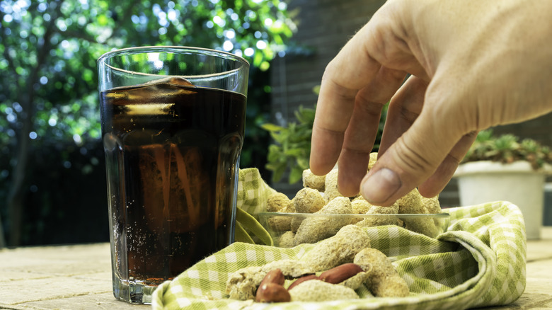A glass of coca cola on picnic table next to bowl of peanuts and hand reaching toward peanuts.