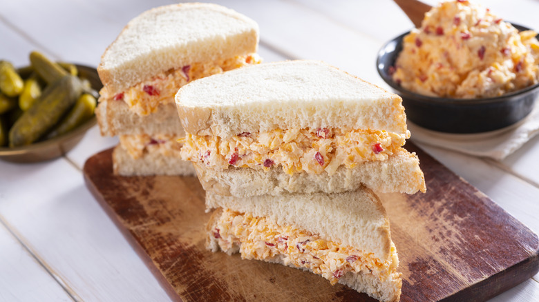 Pimiento cheese sandwiches on a wood cutting board with dish of pickles and mayo salad in background.