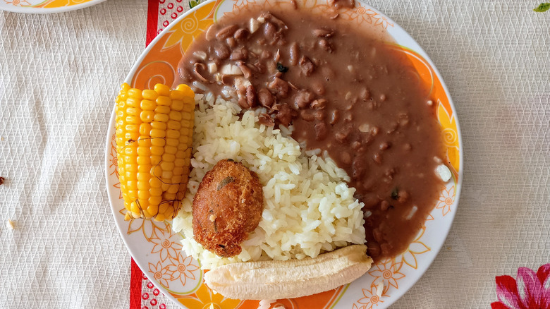A white plate of saucy red beans and rice with corn on the cob and banana on the side