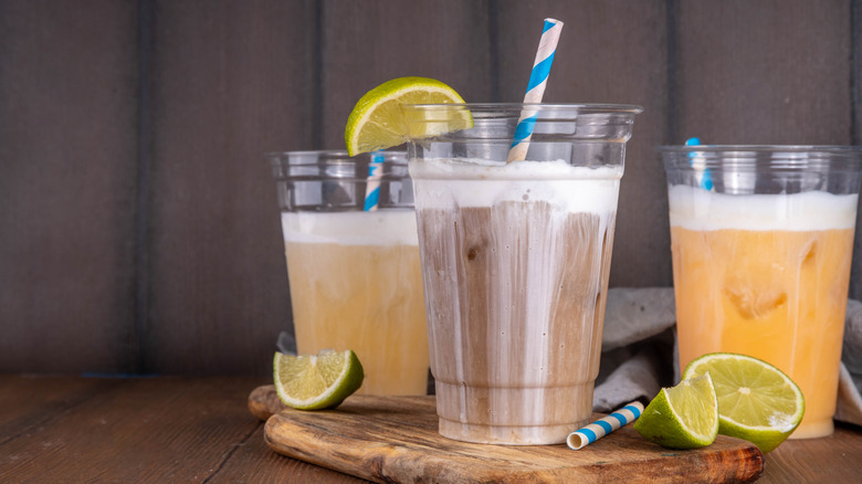 Three clear plastic cups filled with dirty sodas with slices of limes beside them
