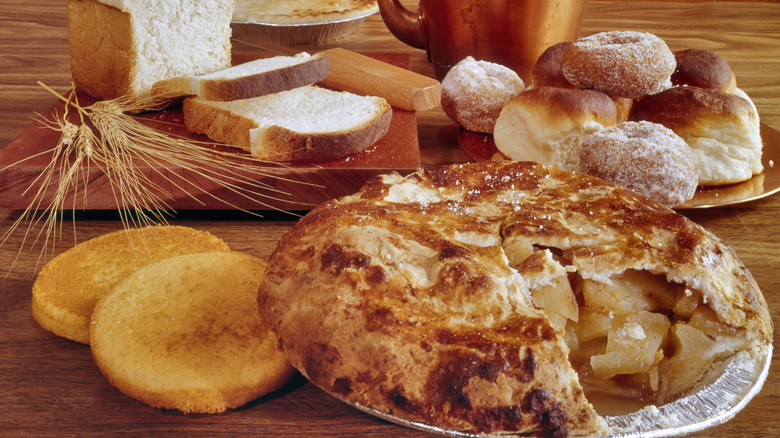 A wood and brass setting with handmade donuts, homemade bread, grains of wheat, and an apple pie