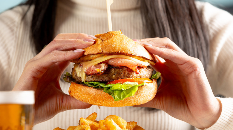 Close up of woman holding a burger prepared to bite into it, over a plate of fries and next to glass of beer