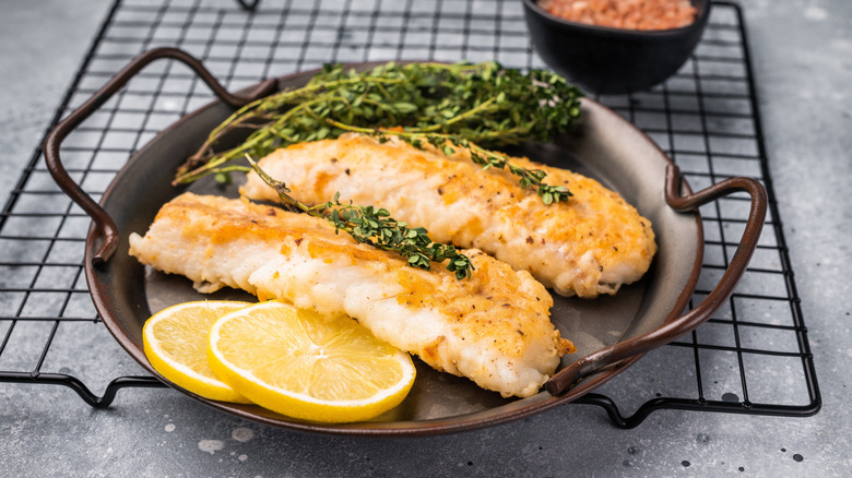 A metal plate with cooked cod, lemons, and herbs on it.
