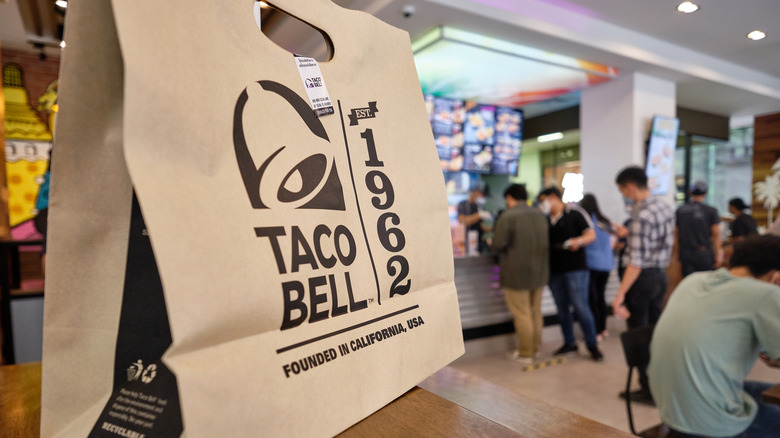 A brown paper bag with the Taco Bell logo on it sitting on the counter in a Taco Bell restaurant with people in the background.
