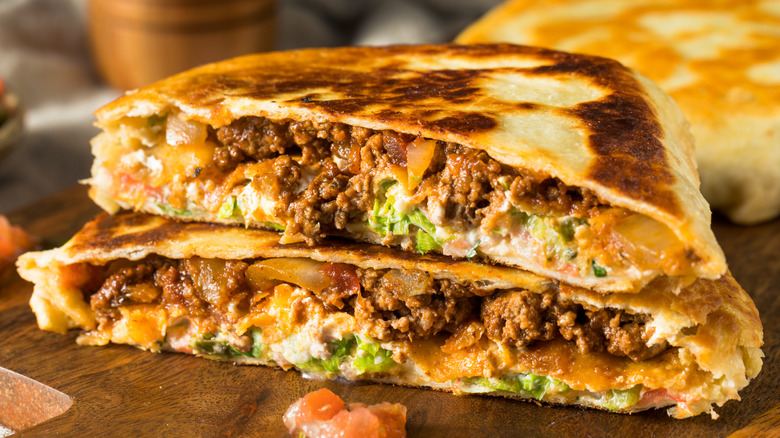 One browned crunchwrap half on top of the other with ground beef, tomato, cheese, and other fillings on a wood surface