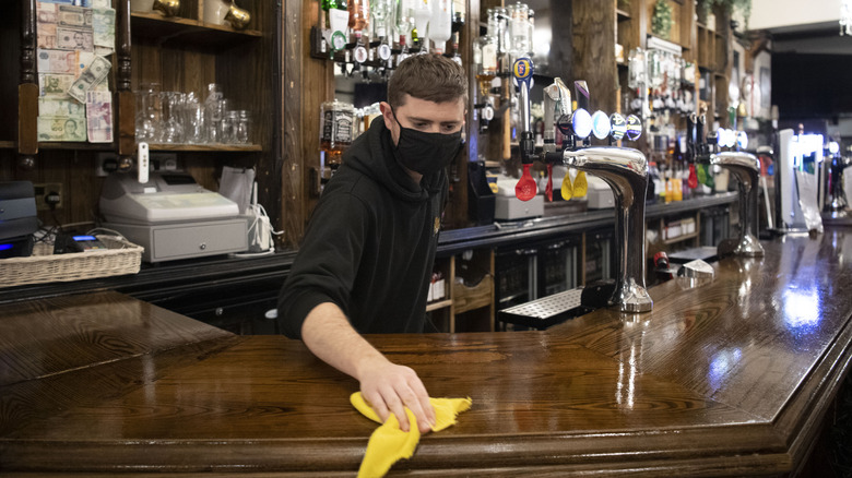 A bartender wearing a mask and cleaning the surface of a bar
