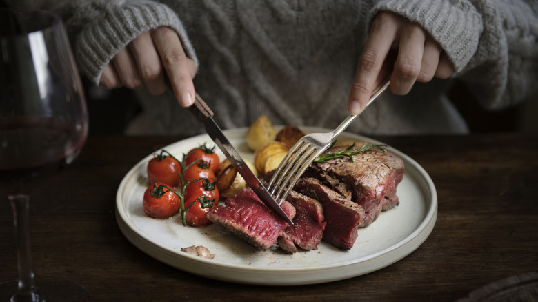 Hands cutting into steak alongside roasted tomatoes and potatoes