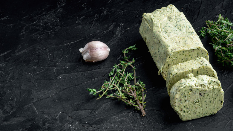 A stick of green garlic butter next to fresh garlic and herbs, on a texturized dark table.