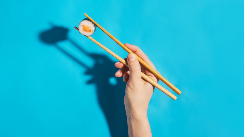 A hand with chopsticks holding a sushi roll against a blue background