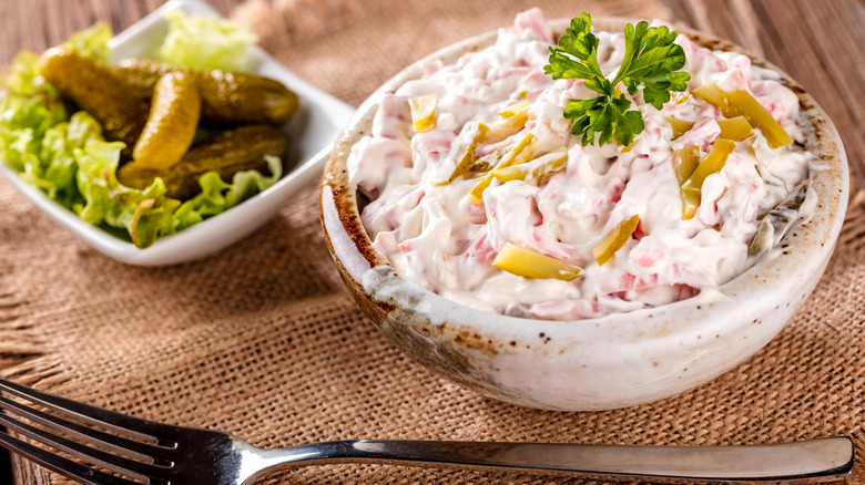 A bowl of bologna salad with mayo on a table next to a dish of small pickles
