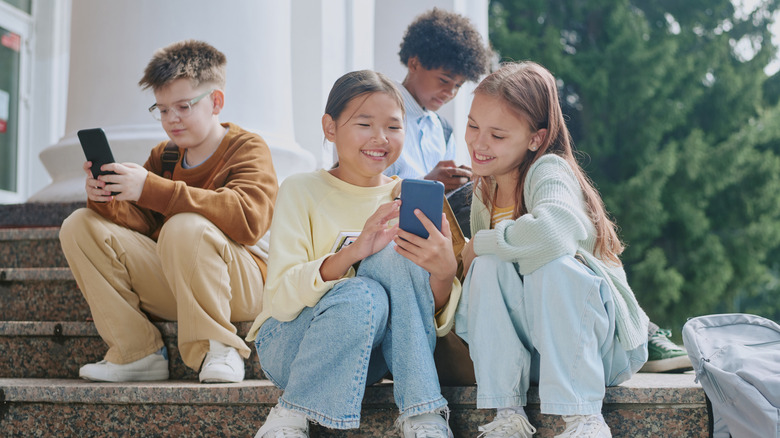 A group of young kids staring and smiling at their phones while sitting on the steps