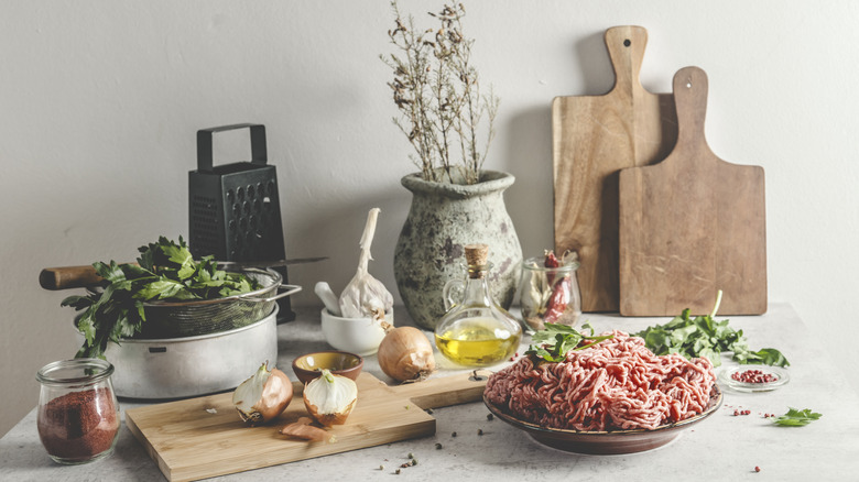 Various ingredients on a marble table with grater, cutting boards, raw meat, onions, oil, and a potted plant.