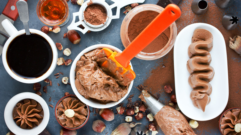 Aerial view of chocolate frostings and spreads with crushed hazelnuts, cocoa powder, coffee, cognac and utensils on a flat surface.