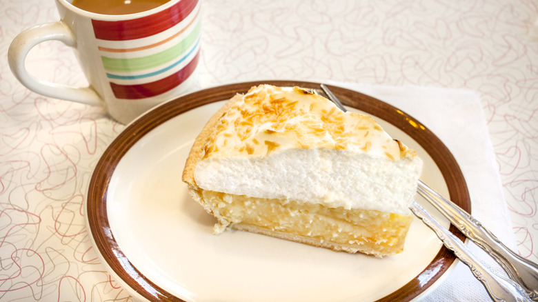 A slice of coconut cream pie on a ceramic plate, served with a mug of coffee