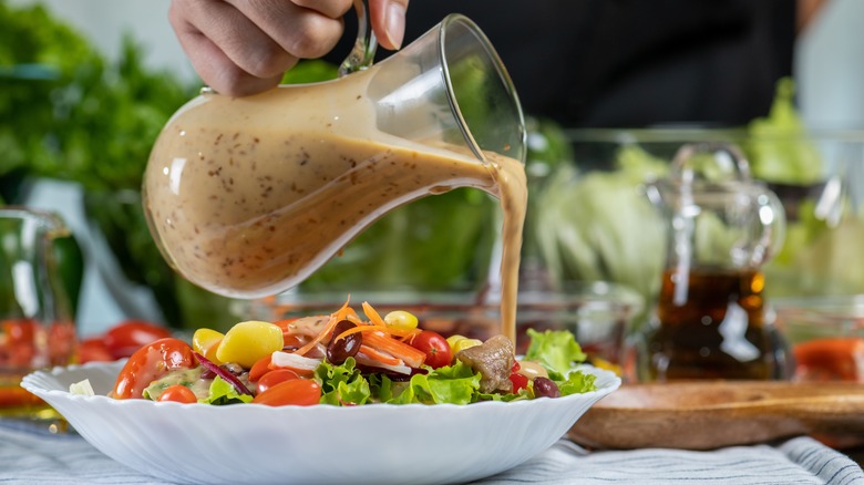 A pitcher of salad dressing being poured over a bowl of salad