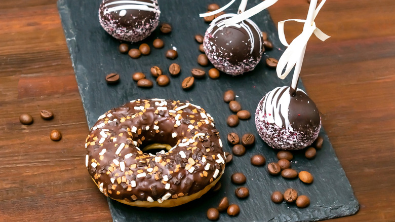 chocolate cake pops decorated with white frosting drizzle on slate board with donut, coffee beans on wooden table