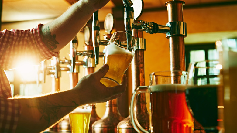 Bartender pouring beer from draft with other types of beer surrounding on a bar top, with pub interior showing in the background during the day.