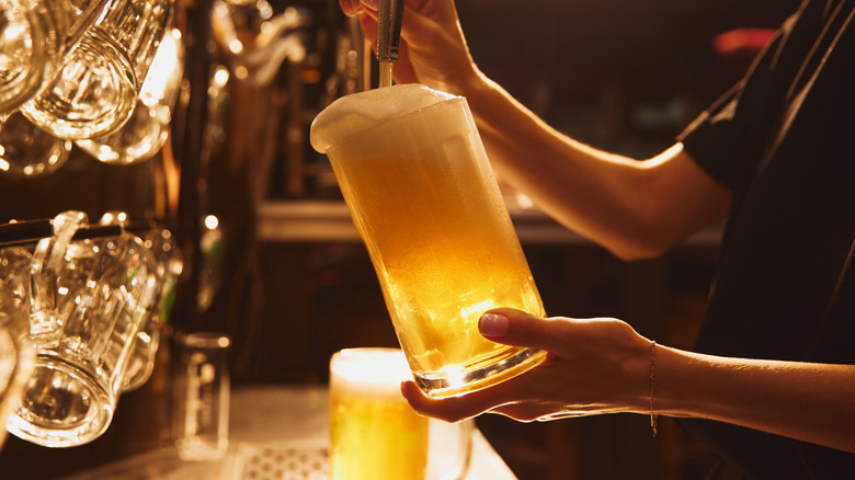 Bartender pouring light beer from a draught into glass mug with empty mugs hanging behind, and a bar setting is in the background.