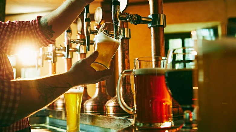 Bartender pouring beer from draft with other types of beer surrounding on a bar top, with pub interior showing in the background during the day.