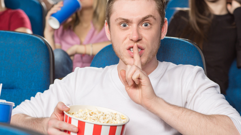 A man shushing someone and holding a bucket of popcorn at a movie theater