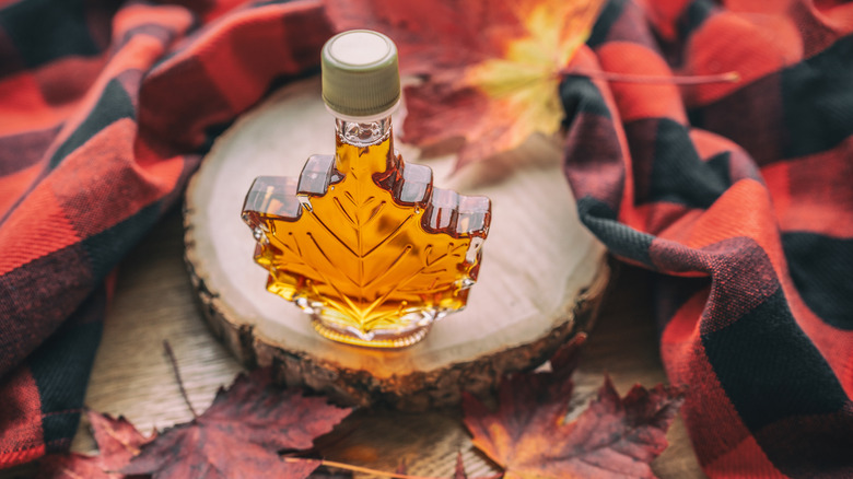 A maple leaf-shaped bottle of maple syrup on a stump surrounded by leaves and flannel