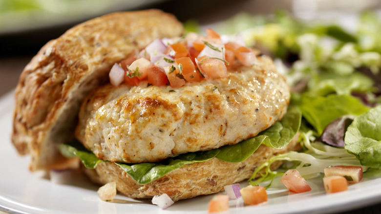 Light-colored turkey burger with chopped tomato and onion on top on an open bun on a white plate with salad greens