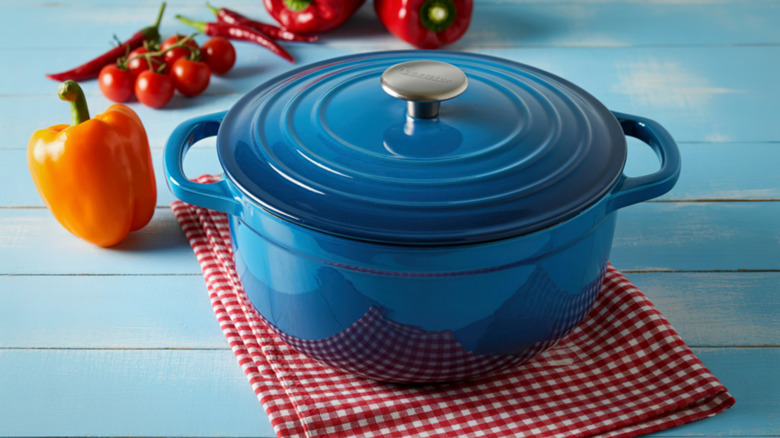 Blue enameled cast iron dutch oven with lid on on top of red gingham kitchen towel with peppers behind it