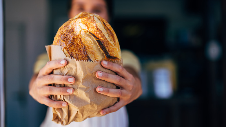 Person's face blurred in the background as they hold a loaf of crusty bread straight ahead, half way wrapped in paper bag