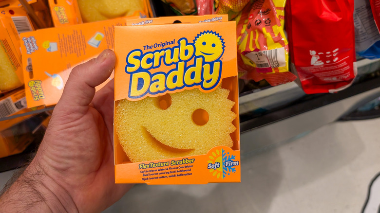 A hand holding a legitimate Scrub Daddy in orange packaging
