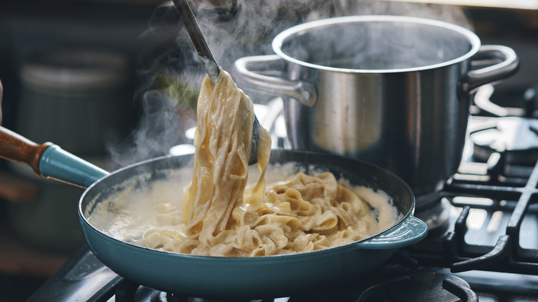 Someone using a utensil to twirl fettuccine Alfredo that's cooking in a blue pan next to a steaming pot.