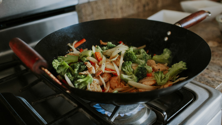 Stir-fry in a wok on a gas stove with broccoli, onions, and chicken.