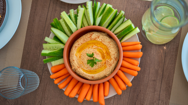 Top view of a bowl of hummus on a plate surrounded by cucumber spears and baby carrots