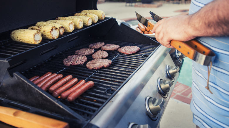 A man holds tongs next to a grill with corn, burgers, and hot dogs