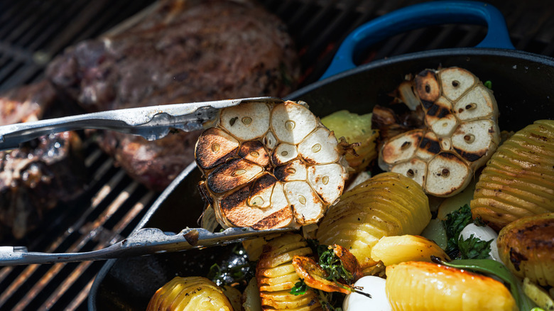 Holding a garlic bulb over food on a grill using tongs