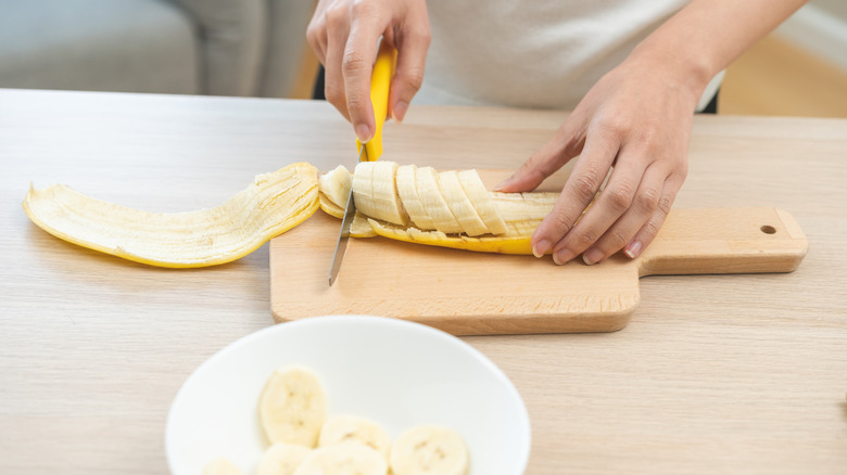 Hands slicing banana on wood board next to white bowl of banana slices