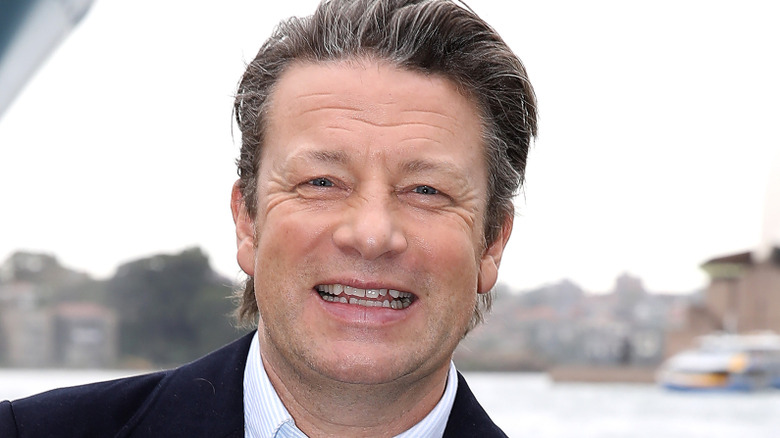 Jamie Oliver smiles in a suit at an outdoor event by the water