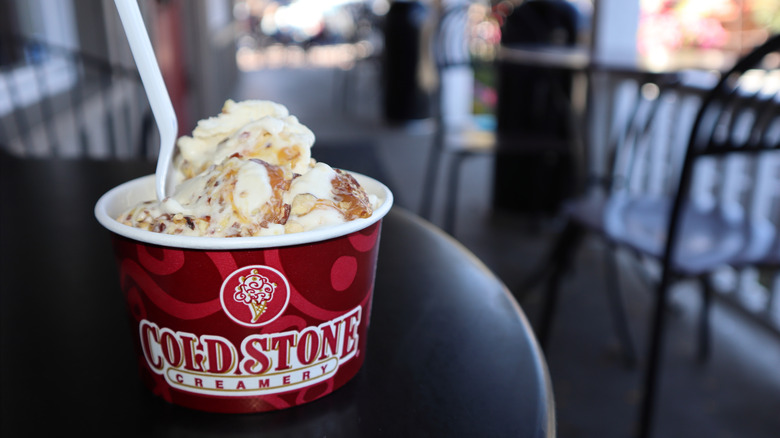 White ice cream with a caramel drizzle in a Cold Stone Creamery cup with a plastic spoon sticking in on a round black table with tables and chairs in the background